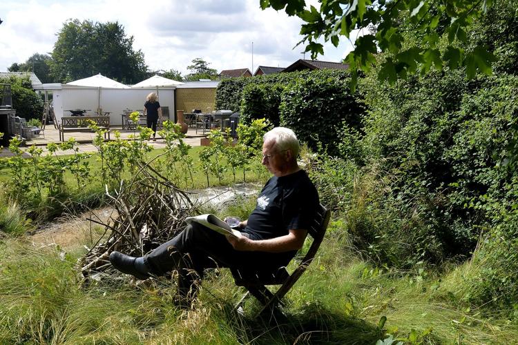 Kurt H. Jørgensen slapper af i sin have med vildt græs, bål og brændestabler og en lille stol. 