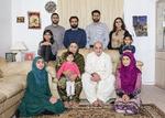 I midten sidder Shamim Hussain med barnebarnet Zainab på skødet og husbonden Munir Hussain ved sin side. Bag dem står parrets børn. Set fra venstre er det Waseem, Mubeen, Faheem og Meena Hussain. På armlænene sidder børnebørnene Dina (tv.) og Sana (tv), på gulvet til venstre barnebarnet Isma og til højre svigerdatteren Hafsa. 