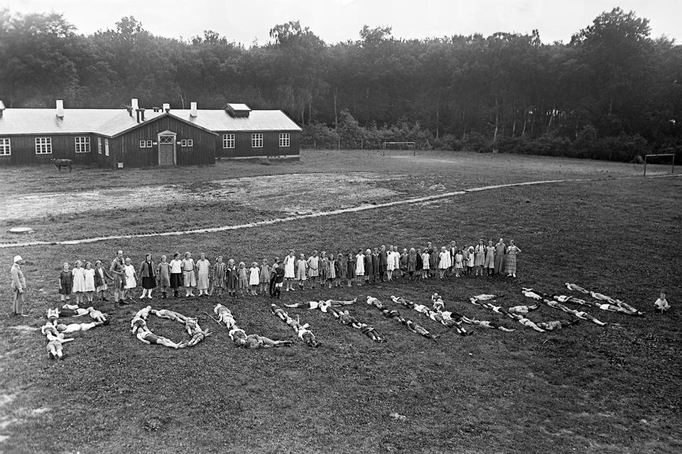 Den årlige indsamling blandt Politikens læsere har i sommeren 1923 gjort det muligt at sende nogle af storbyens fattige børn på feriekoloni i Horserødlejren. De taknemlige piger har skrevet ’Politiken’ med sig selv som bogstaver til ære for fotograf Holger Damgaard og Politikens læsere. Bemærk i øvrigt, at lejren har egen ko.  Foto: Holger Damgaard