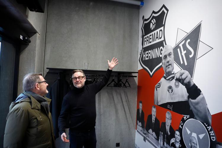 Indgangspartiet i FC Midtjyllands nye hovedkvarter, Dream99, er udsmykket med en kollage over milepæle i klubbens historie. Formand Claus Steinlein udpeger her for Helge Sander, at den tidligere Venstre-minister er med og gemmer sig bag Herning Fremads logo.  Foto: Jens Dresling