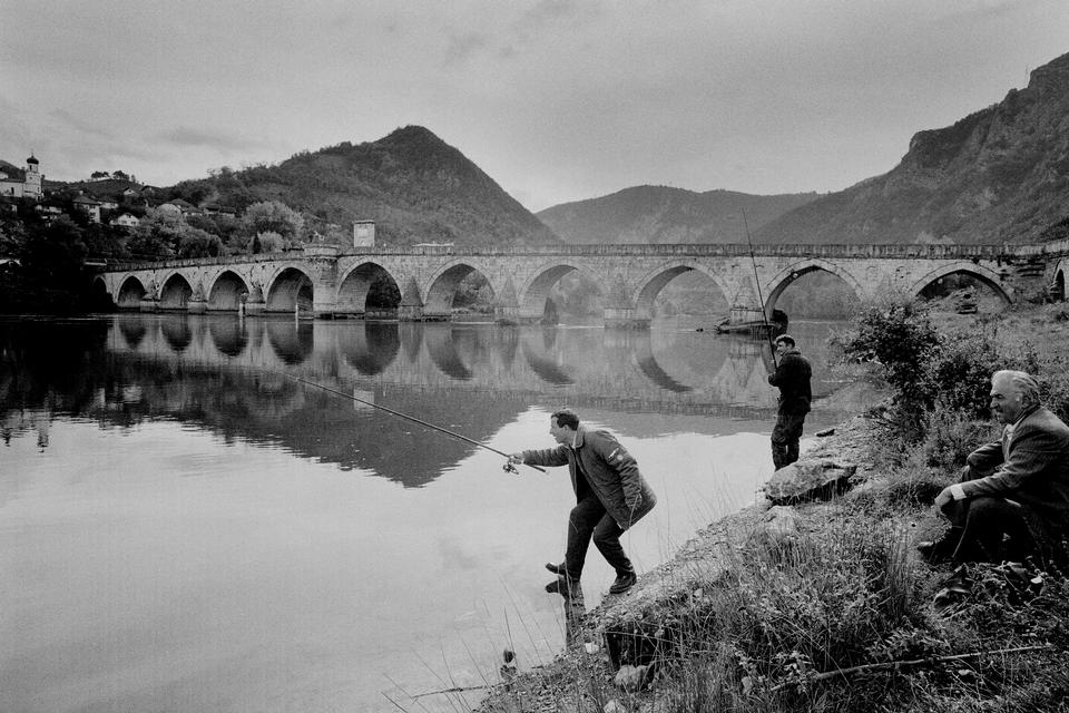 (Visegrad, Bosnien, 1994).  »Nede ved Drina-floden fiskede nogle mænd i et landskab, der også bar sporene af krig. Over dem spændte den gamle bro, kendt fra Ivo Andrics roman Broen over Drina, et sted, der i sig selv fortæller om mødet mellem kulturer og brudflader i historien. I det stille øjeblik ved vandet var der både ro og en underliggende spænding i tiden«.  Foto: Carsten Ingemann/INGEMANN PHOTOGRAPHY