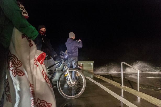 Godt pakket ind i vinterjakker, huer og tæpper er folk taget til Amager Strandpark, hvor de her står tæt på bølgerne, der ved hårde vindstød sprøjter højt op over deres kroppe.

 