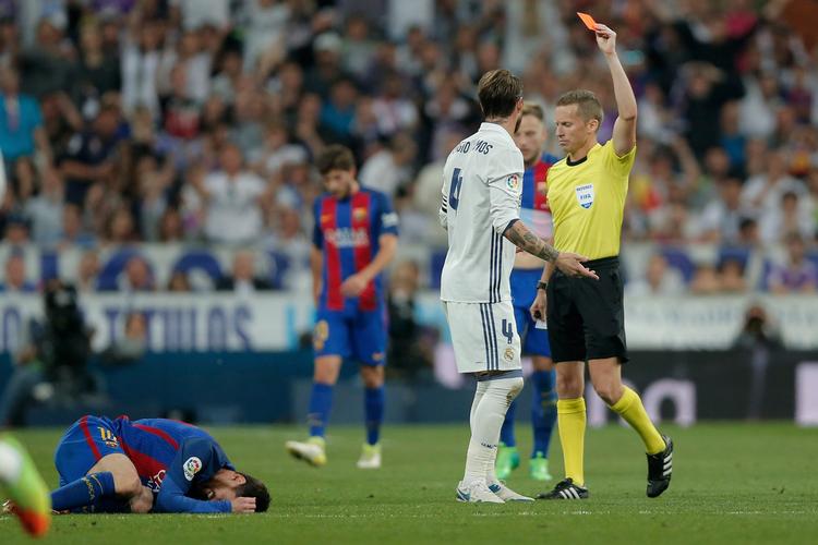 Real Madrids anfører Sergio Ramos får marchordre efter sin vilde tackling, mens Barcelonas Lionel Messi gør, hvad alle angribere gør: spiller rollen som død.  