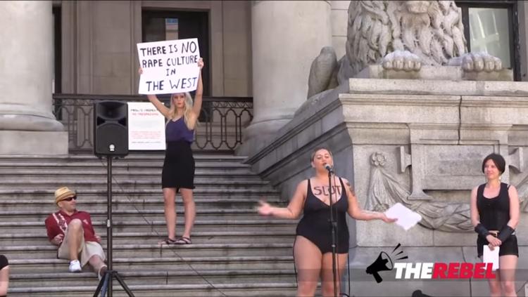 Lauren Southern holder et skilt med et provokerende udsagn i vejret bag taleren ved en feministisk demonstration i Vancouver, Canada, i juni 2015.  Therebel media