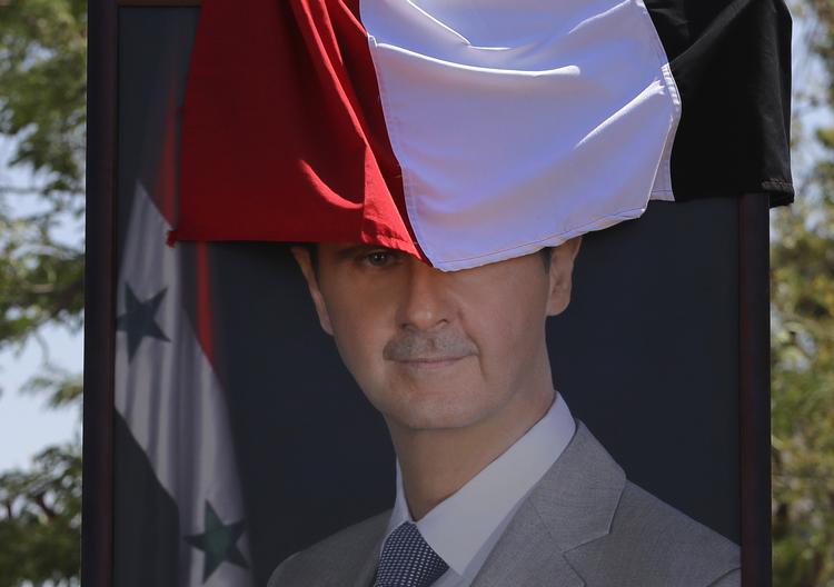 Et billede af den syriske præsident, Bashar Assad, er halvt dækket af det syriske flag. Diktatoren soler sig i, at hans militær – med hjælp fra bl.a. Rusland og Iran er ved at genvinde magten i langt det meste af landet. Foto: Sergei Grits/AP