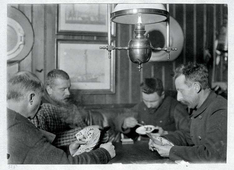 Til trods for en del drama undervejs, havde deltagerne på Danmark-ekspeditionen også mange hyggelige stunder med musik og bøger og kortspil.  Her er det fra venstre læge Johannes Lindhard, premierløjtnant og kartograf J.P. Koch og ornitolog Arner Manniche, der sidder og spiller kort, mens maleren og forfatteren Achton Friis spiser 