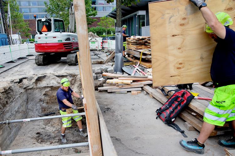 Solen bager ned over København, mens Kenneth Most er i gang i fjernvarmeudgravning. Tom Larsen i baggrunden  tilpasser planker, og Henrik Jørgensen slæber en krydsfinerplade.   Foto: Finn Frandsen/POLFOTO