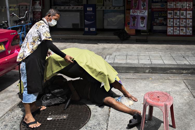 Denne mand faldt torsdag om på en gade midt i Bangkok. Det er uvist, om årsagen var coronarelateret. I flere timer prøvede forbipasserende at tilkalde en ambulance, men kunne ikke komme igennem til alarmcentralerne, der med flere tusinde coronaopkald om dagen er lagt ned. I stedet forsøgte beboere i gaden at mindske mandens lidelser en smule ved at skærme ham for den skarpe sol. Foto: Claus Blok Thomsen
