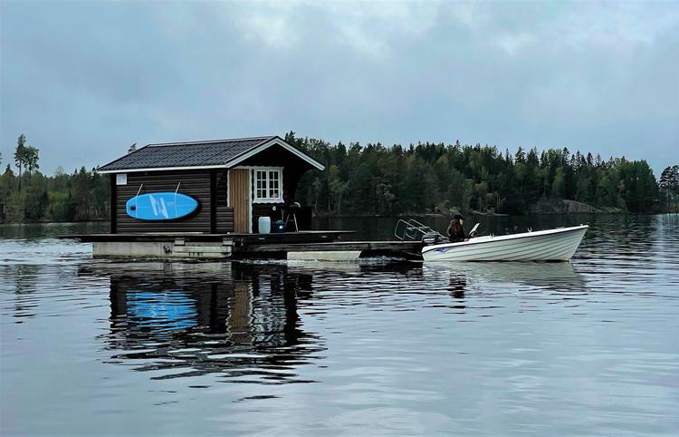 Det lille træhus er placeret på tømmerflåden, som den hvide motorbåd sejler rundt på søen. Midt ude på søen kan du fiske, stå på SUP, bade, spotte elge eller bare kigge ud på det blikstille vand.  Foto: Eva Holtegaard-Kasler
