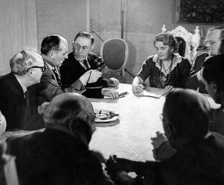 Spiritisten Anna Melloni Rasmussen afsløres i falsk åndemaneri. Fra venstre: redaktør Karl Bjarnhof, redaktør Dahlerup, ægteparret Melloni, prof. Plum, direktør Peter Linholt Nielsen, forfatter Jacob Paludan og kunsthistoriker Aage Marcus. På det andet foto ses det hul, der blev banket i væggen, så fotografen kunne optage, hvad der skete under bordet. Foto: Jacob Maarbjerg
