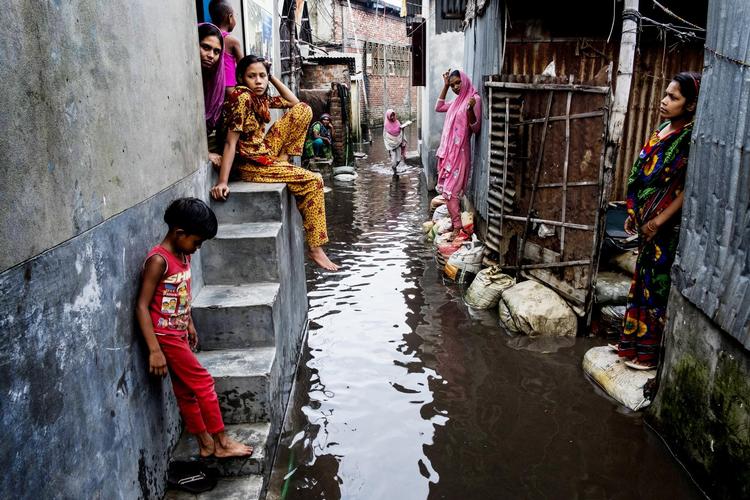 Når folk ude på landet opgiver kampen mod fattigdom, naturkatastrofer og klimaforandringer, flytter de til store byer for at søge arbejde. De fleste ender i Dhakas slum. Her begynder mange at gruble over, om de mon kan finde en vej til udlandet. Kvinderne på billedet nyder et kort ophold i monsunregnen i slumkvarteret, der kaldes Tændstikkolonien. 