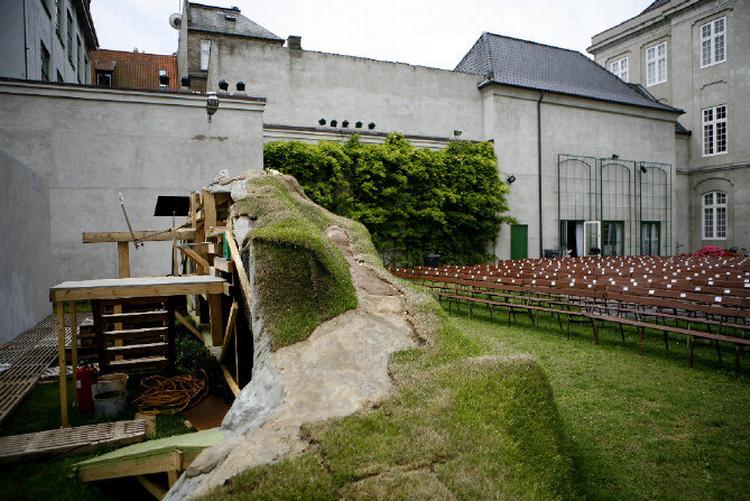 Til opsætningen af 'Glade dage' er der skabt en sindrig scenografi, hvor skuespilleren Kirsten Lehfeldt kan hejses op og ned i sin jordhøj. Til højre ses resterne af Odd Fellow Palæet, som blev bygget 1751-55 af gehejmeråd August von Berckentin. Fra 1889 rummede bygningen også en stor koncertsal. 