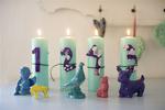BARNLIG ADVENTSKRANS. Fire mintgrønne bloklys stillet på række med tal af tapeter bundet omkring lysene med en sort snor. Porcelænsdyr fundet i en genbrugsbutik er spraymalet i forskellige farver. 