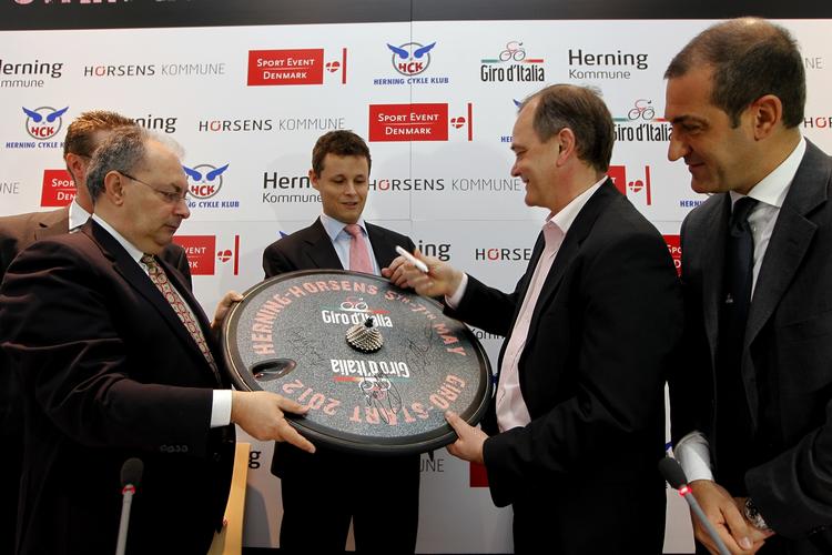 giro. De to borgmestre fra Herning og Horsens, Lars Krarup og Jan Trøjborg, modtager en lille Giro-markering fra Giro d'Italias direktør Giacomo Catano (th). 