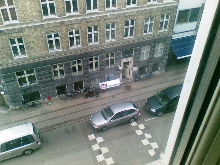 Bedemanden venter ved den åbne kiste med den afdøde kvinde, mens en anden bedemand er oppe i kvindens lejlighed for at hente de pårørende. 