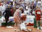 knivstik. Monica Seles sidder på gruset efter angrebet, mens sikkerhedsfolk pågriber knivstikkeren på tilskuerrækkerne. 