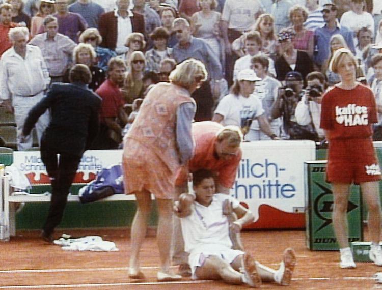 knivstik. Monica Seles sidder på gruset efter angrebet, mens sikkerhedsfolk pågriber knivstikkeren på tilskuerrækkerne. 