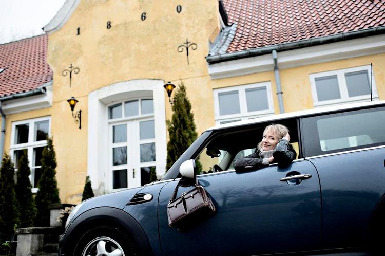 Præsteboligen. Her er Kathrine foran præsteboligen som kom med embedet iført hendes 18 år gamle Kenzo jakke, med hendes slangeskindstaske købt for 50 kroner over armen, ved siden af hendes Mini Cooper, der blev købt på fem minutter. 