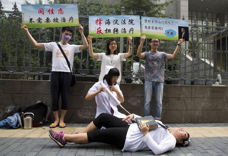 PROTEST. »Homoseksualitet er ikke en sygdom. Vi behøver ingen kur«, står der på skiltene, som holdes af en gruppe aktivister uden for Beijings retsbygning i dag.  Nederst foregiver den lokale LGBT-formand, Xin Ying at være en læge, der giver en anden aktivist elektrochok-terapi. 