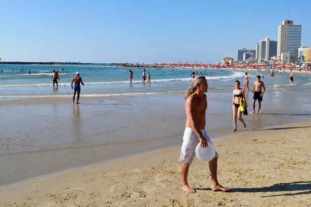 Jerusalem Beach. Et fluepapir med parasoller, liggestole og folk, der bader og spiller strandtennis. I vandkanten er der lidt plads til at spille frisbee. 