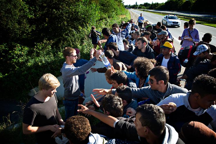 Ved et hul i de store buske og træer, der er langs motorvejen, har tre unge fyre stillet sig op på en lille afsats, og giver flygtningene mad og vand. Flygtningegruppen flår maden til sig, og det er tydeligt, at de er sultne og trætte. 