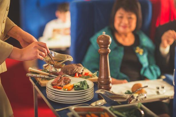 Stewardessen skærer bøffen til ved den rejsendes sæde. Alt tilbehør er også på plads. 